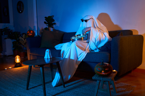 Sheet ghost sits on a couch, eating popcorn and watching a movie. 